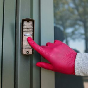 A red gloved hand presses a modern doorbell on a metal door, depicting security and service.